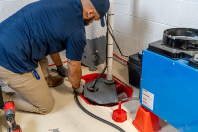 A person in a blue shirt expertly installs a PVC pipe onto a blue and red sump pump on gravel, ensuring optimal drainage for effective basement waterproofing.