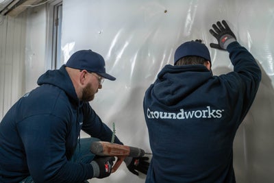 Two men install a vapor barrier onto a basement wall with tools and protective gear.