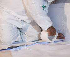 Person in protective suit taping plastic sheet to floor with white tape.