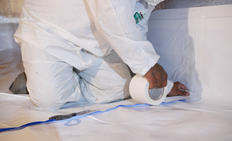 Person in protective suit taping plastic sheet to floor with white tape.