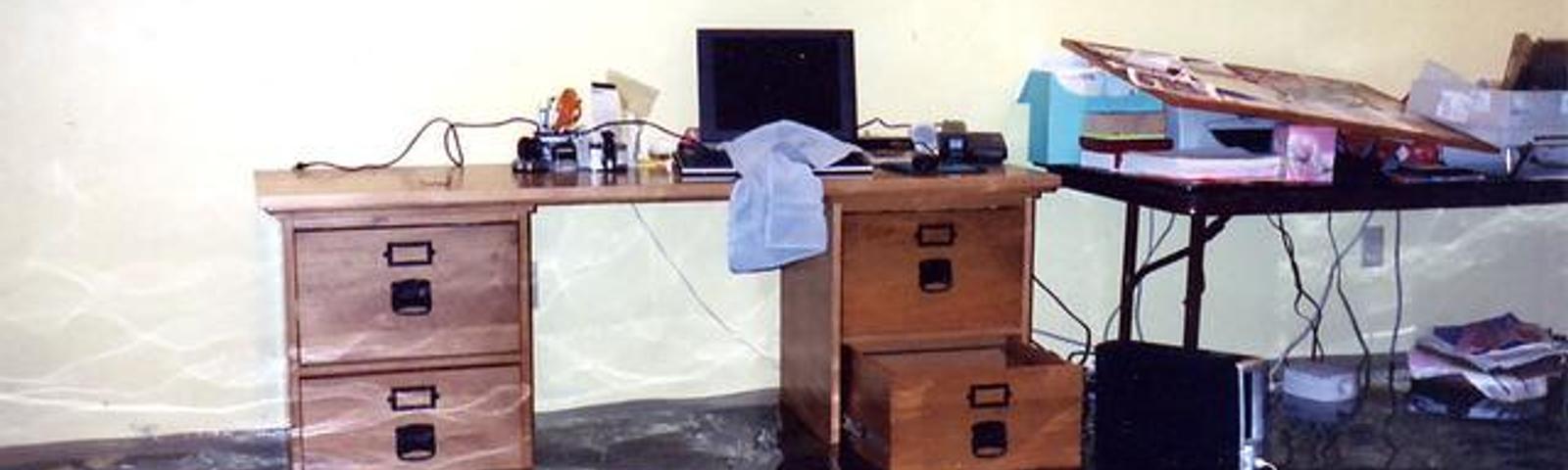 A flooded room with a wooden desk and a table, both partly submerged in water.