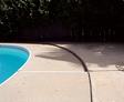 Curved edge of a swimming pool with a concrete deck and a wooden fence in the background.