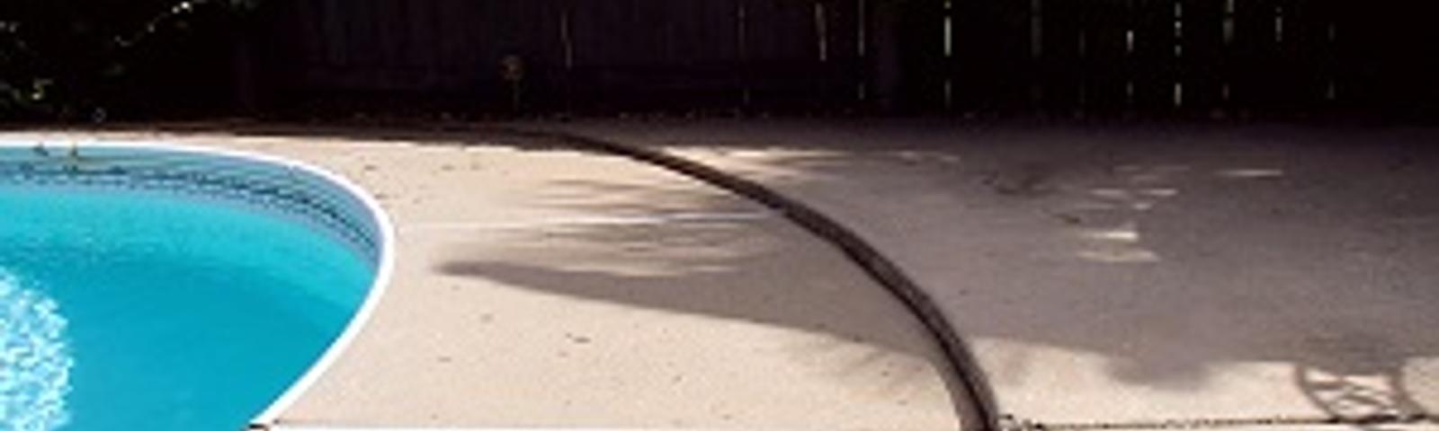 Curved edge of a swimming pool with a concrete deck and a wooden fence in the background.