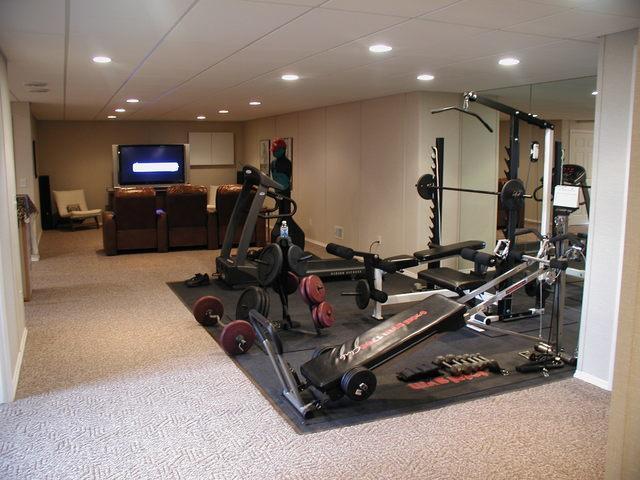 Home gym with various exercise equipment, mirrored wall, and nearby seating area with a large TV screen.