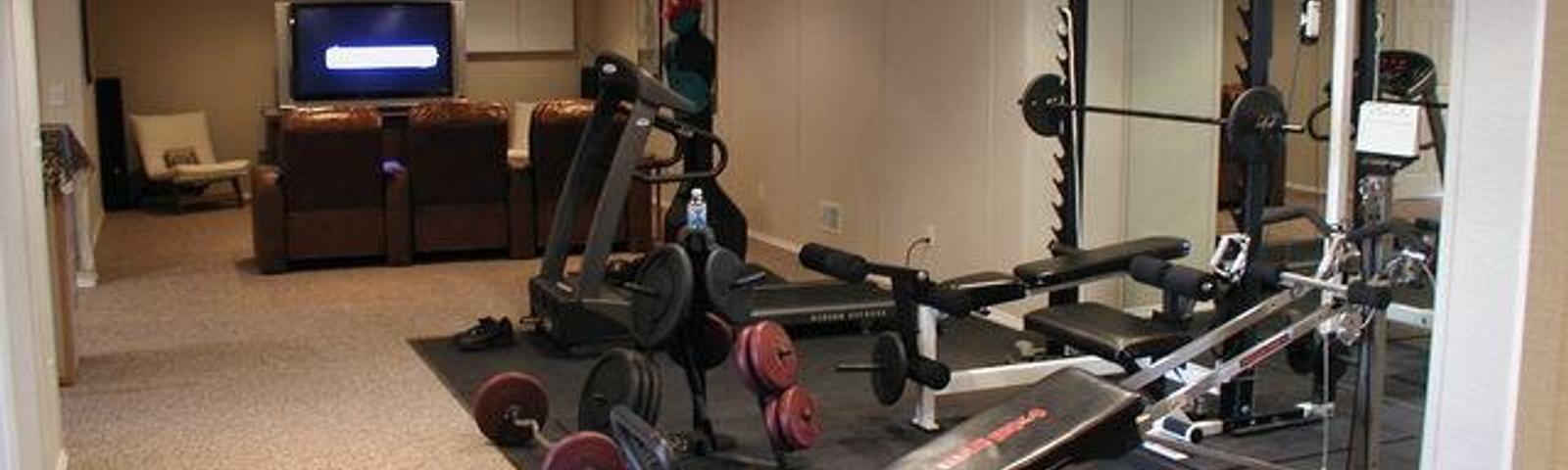 Home gym with various exercise equipment, mirrored wall, and nearby seating area with a large TV screen.