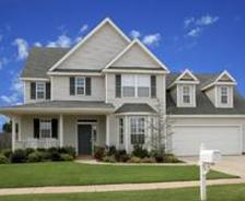 A two-story house with a gray roof, front lawn, and attached garage under a blue sky.