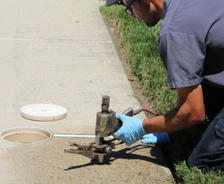A worker in blue gloves repairs a water valve on a sidewalk near a patch of grass.