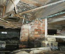 View of a damp, deteriorating crawl space with exposed wooden beams and brick supports.