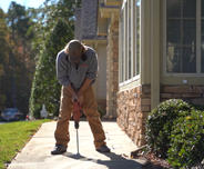 Person using a jackhammer on a sidewalk next to a brick house, with trees and grass in the background.