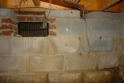 A basement wall with exposed cinder blocks and a vent, featuring some hanging wires under wooden ceiling beams.