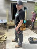 Man in work attire with tools stands outside a building under construction. Another person is in the background.