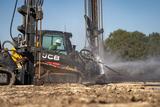 Construction worker operating machinery to spray water on dirt, with trees in the background.