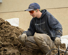 Worker in a blue cap and protective gear kneels by a pile of dirt, holding a tool with focus on the task.
