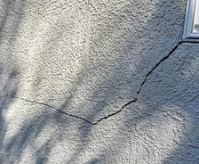 Cracks on a gray stucco exterior wall near a window, with shadows cast on the surface.