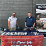 Two men stand behind a table with sunglasses at an Innovative Basement Authority booth, smiling at the camera.