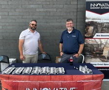 Two men stand behind a table with sunglasses at an Innovative Basement Authority event booth.