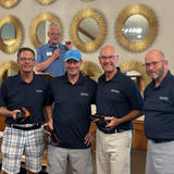 Four smiling men in matching navy shirts hold prizes, standing in front of a man speaking at a podium.