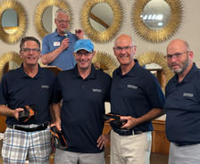Four smiling men in matching navy shirts hold prizes, standing in front of a man speaking at a podium.