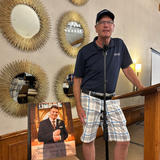 Man in a blue shirt and plaid shorts speaks at a podium next to a memorial photo of another man.