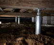 Crawl space with steel support jacks beneath a wooden structure, on a soil floor.