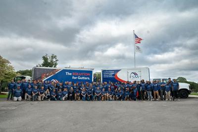 A large group of people in blue shirts stands in front of trucks and an American flag outdoors.