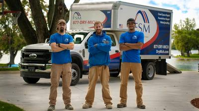 Three workers in blue FSM shirts stand in front of a company truck on a driveway near a lake.