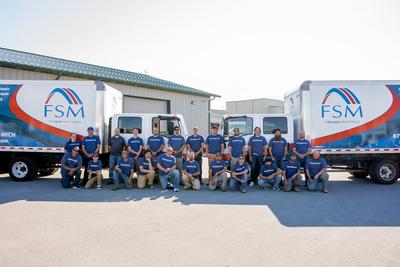 A group of workers in blue shirts pose in front of two FSM company trucks outside a building.