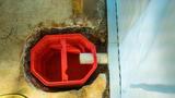 A red sump pump basin, part of a basement drainage system, is set in concrete with a white pipe connected.