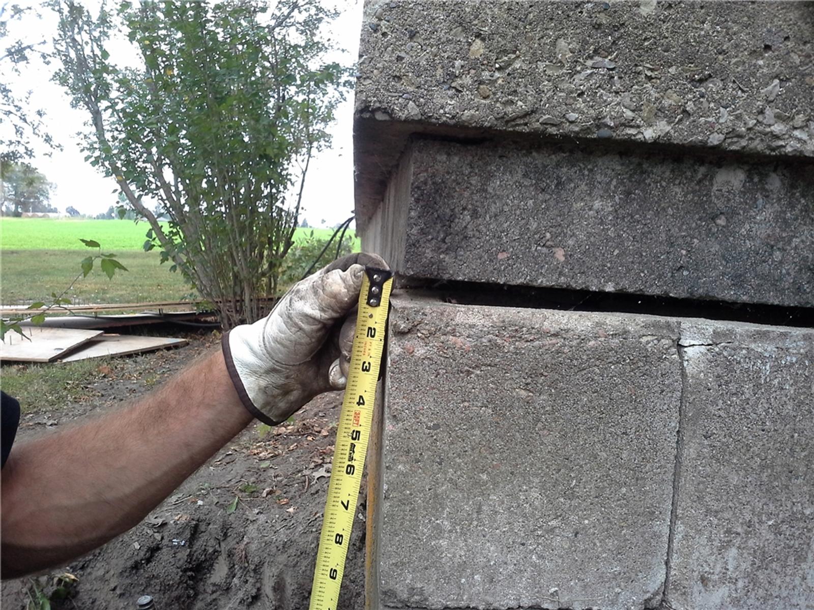 Gloved hand holding a tape measure against a concrete structure outdoors. Trees and grass are visible in the background.