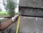 Gloved hand holding a tape measure against a concrete structure outdoors. Trees and grass are visible in the background.