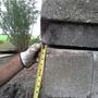 Gloved hand holding a tape measure against a concrete structure outdoors. Trees and grass are visible in the background.
