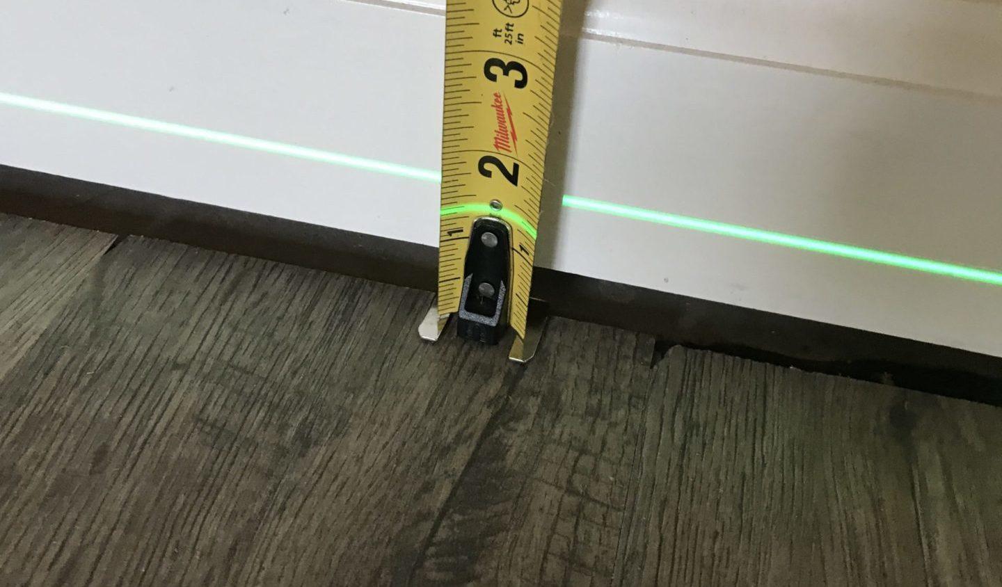 Tape measure against a white baseboard and a green laser level line above wood flooring.