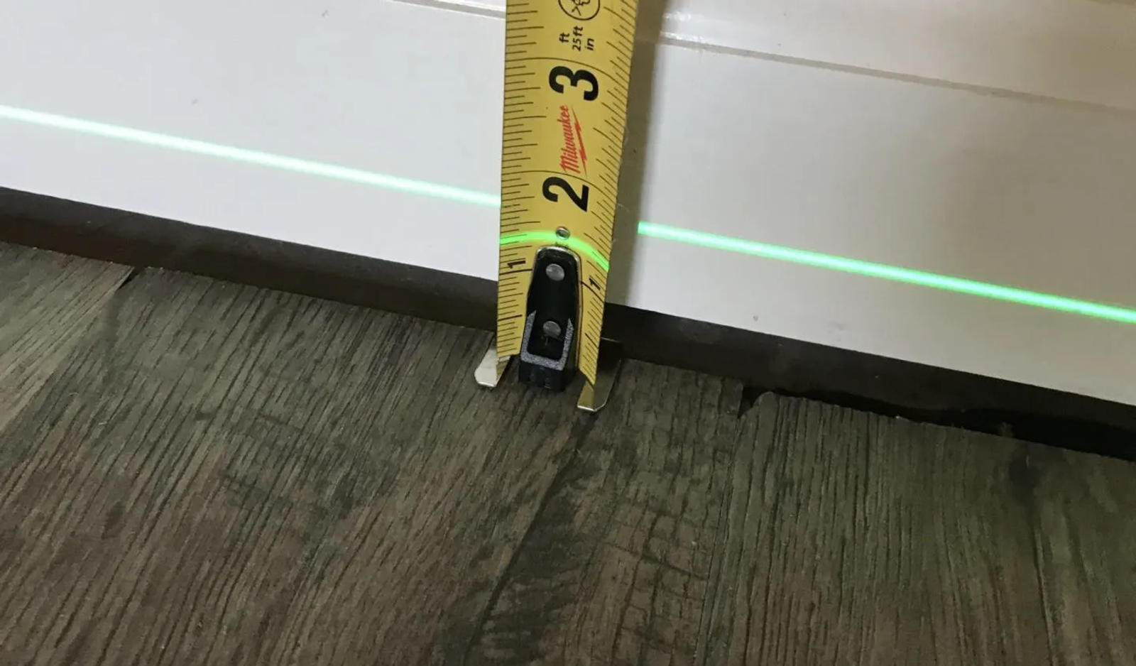 Tape measure against a white baseboard and a green laser level line above wood flooring.