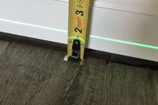 Tape measure against a white baseboard and a green laser level line above wood flooring.