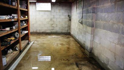 Wet basement with concrete walls, a puddle on the floor, and shelves filled with various tools and items on the left.