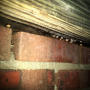 Water droplets hanging from wooden beams above red brick wall, illuminated in dim lighting.