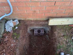 A brick wall with an exposed foundation hole, soil, and bricks scattered around. A metal block lies in the hole.