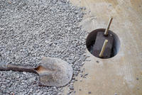 A shovel beside gravel and a concrete surface with a circular hole showing metal rods inside.