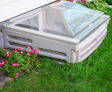 A plastic window well cover next to a house surrounded by grass and pink flowers.