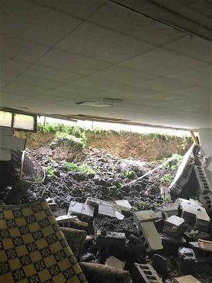 A collapsed ceiling with debris and mud fills the room, revealing sunlight and grass through the opening. The chaos mirrors the bowing basement wall below, suggesting further structural concerns that invite nature inside.