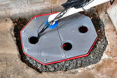 Basement floor with a red-bordered sump pump pit cover, fitted with white and black pipes.