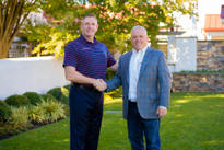 Two men shaking hands and smiling in a garden, standing on a lawn with trees and a stone wall in the background.