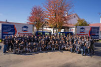 A large group of people in matching shirts pose in front of two BQ Basements trucks with a backdrop of autumn trees.