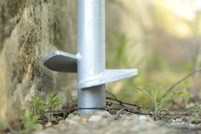 Metal post with angled footplates secured in the ground, surrounded by small plants and gravel.