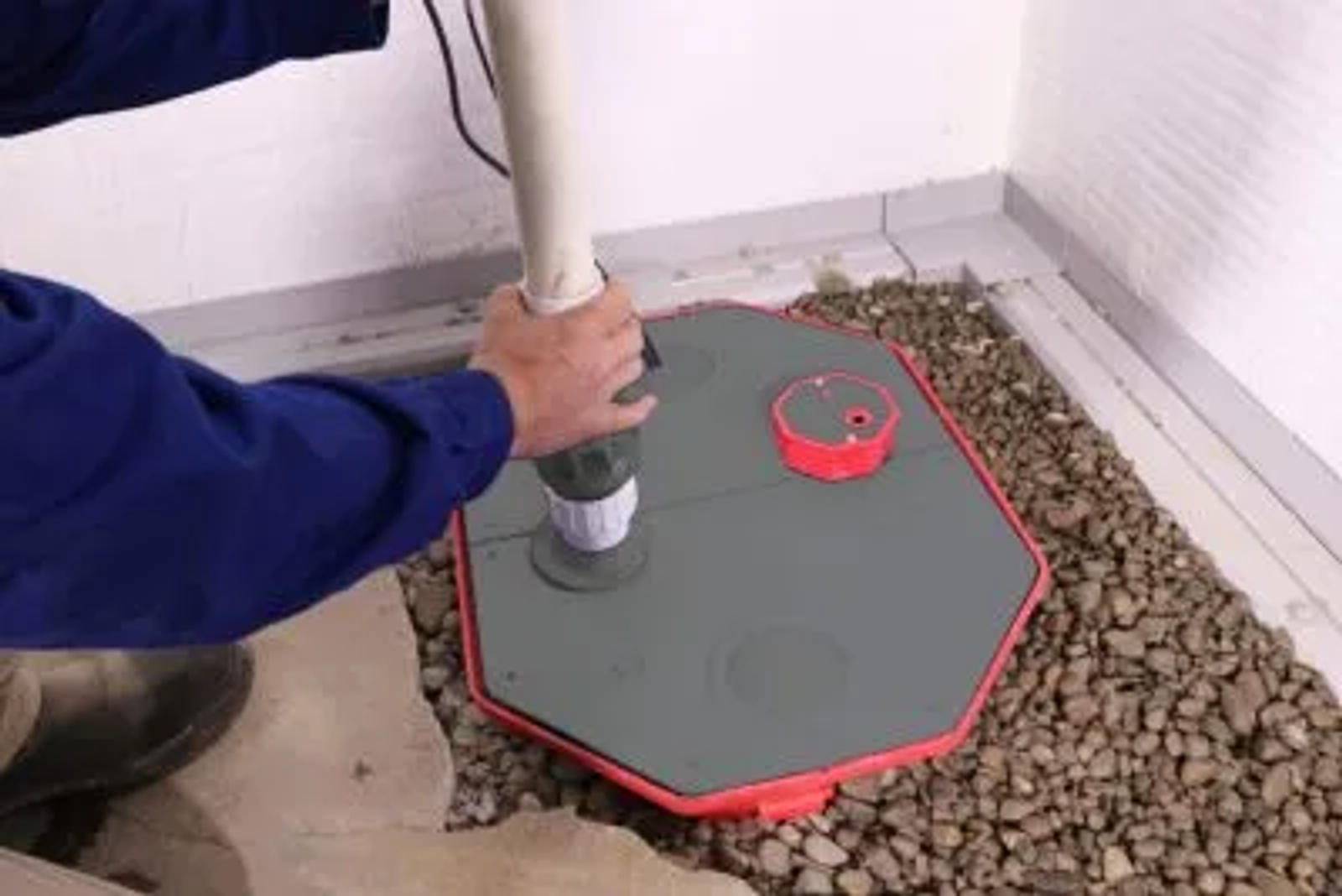 Person installing a drainage system in a rocky area indoors, securing a pipe into a covered basin.