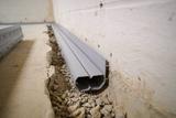 Close-up of gray plastic ducting installed along a concrete floor with exposed gravel in a basement.