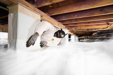 Worker in protective gear insulating a crawl space under a wooden floor using white plastic sheeting.