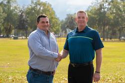 Two men shaking hands in a park, wearing casual clothing, with trees in the background.