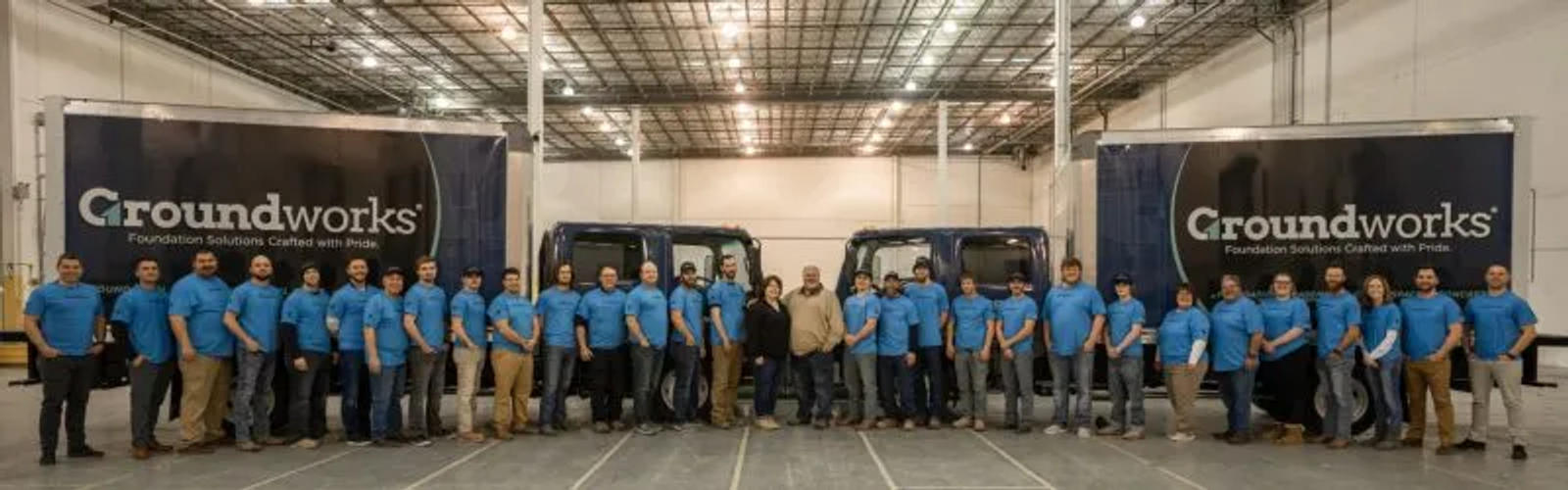 A group of people in matching blue shirts stand in front of trucks inside a warehouse.