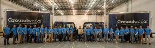 A group of people in matching blue shirts stand in front of trucks inside a warehouse.
